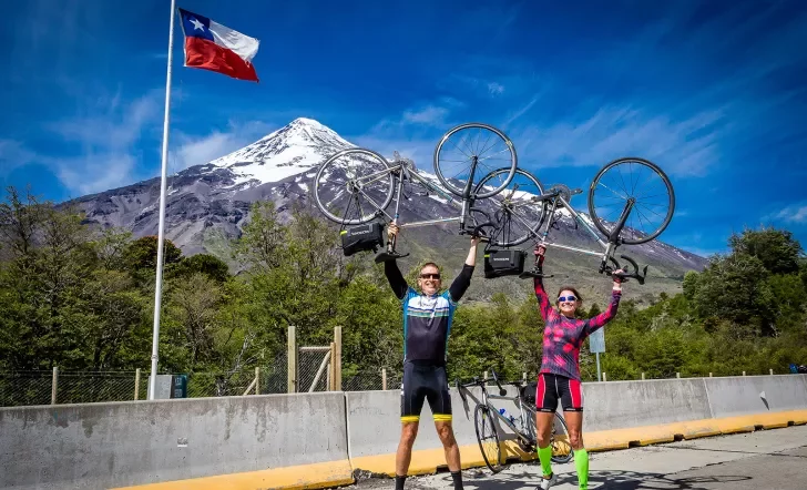 Two guests on road, holding bikes over head, Chilean flag, mountain behind them.