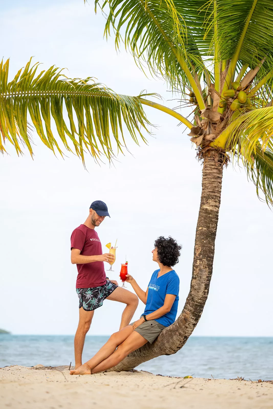 Man and women holding drinks while sitting on a slanted tree on the beach