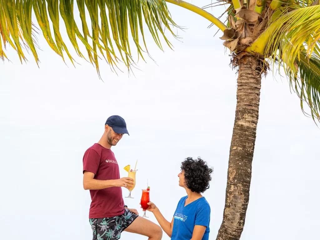 Man and women holding drinks while sitting on a slanted tree on the beach
