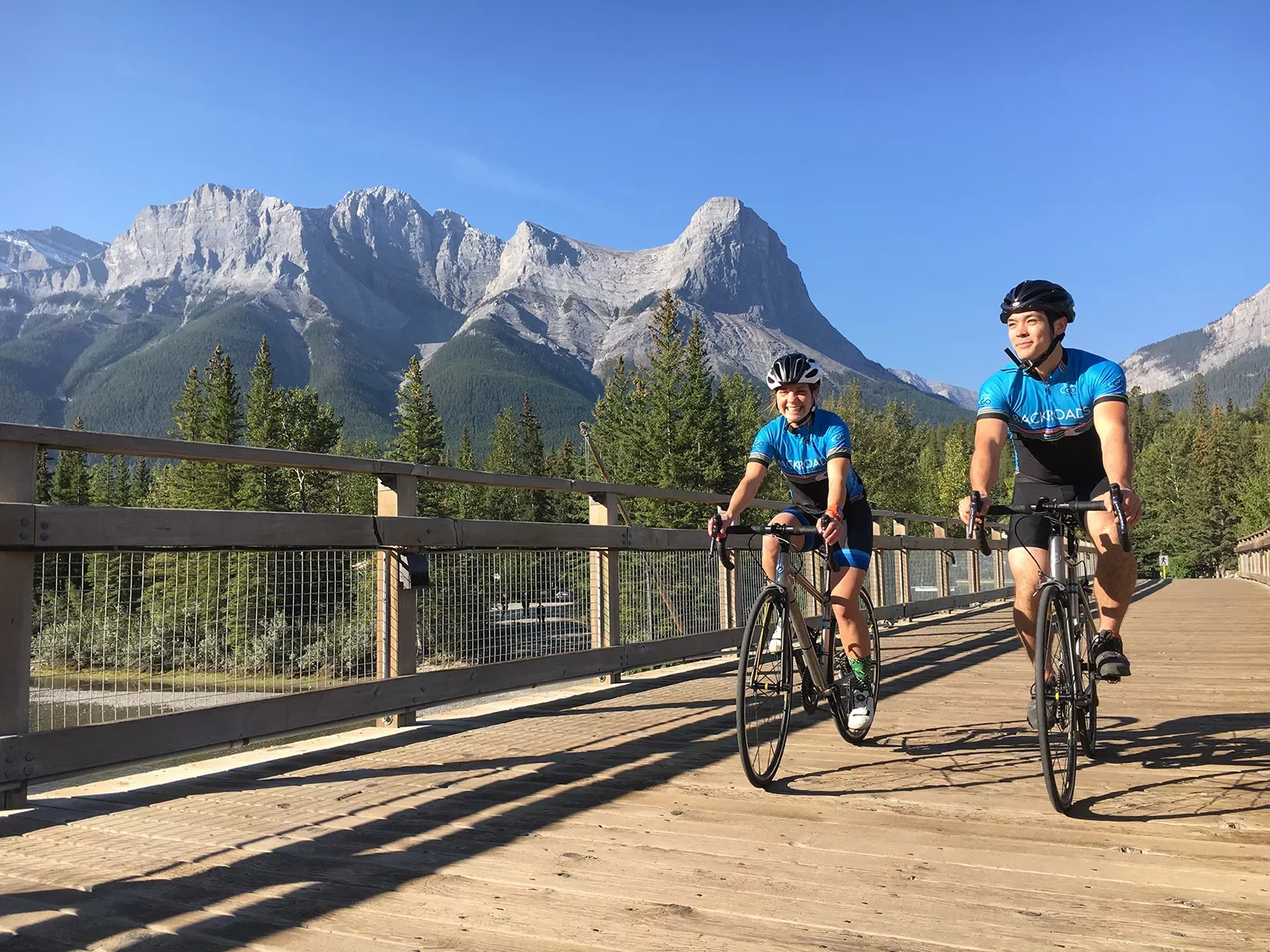 Two guests cycling on wooden path, Rockies in distance.