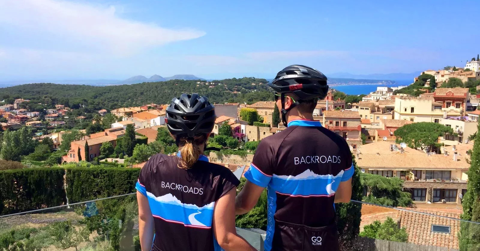 Two guests in bike gear overlooking countryside, tan stone houses, trees all around.