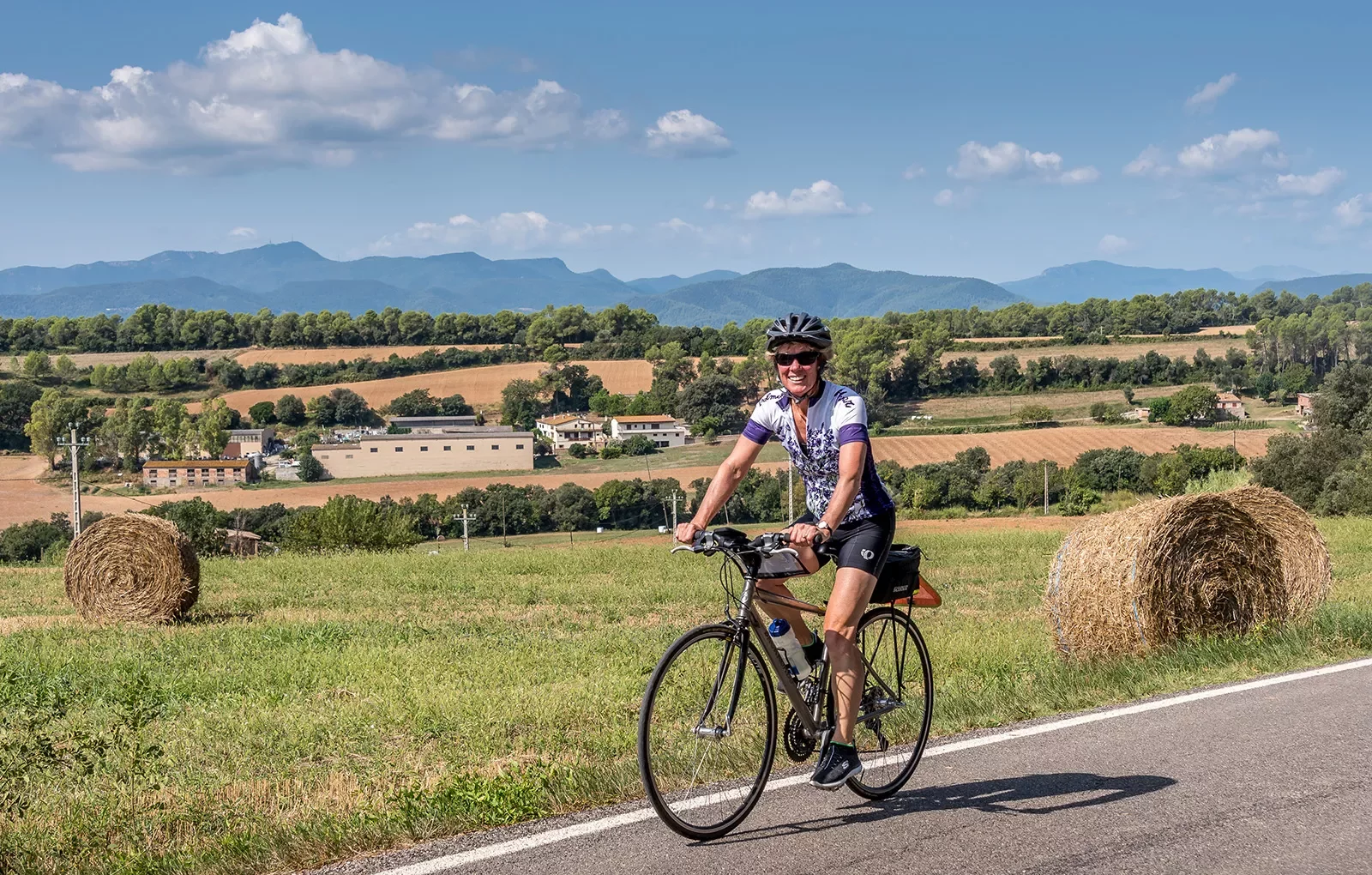 Guest cycling past farmland, hay bales, hilly vista behind her.