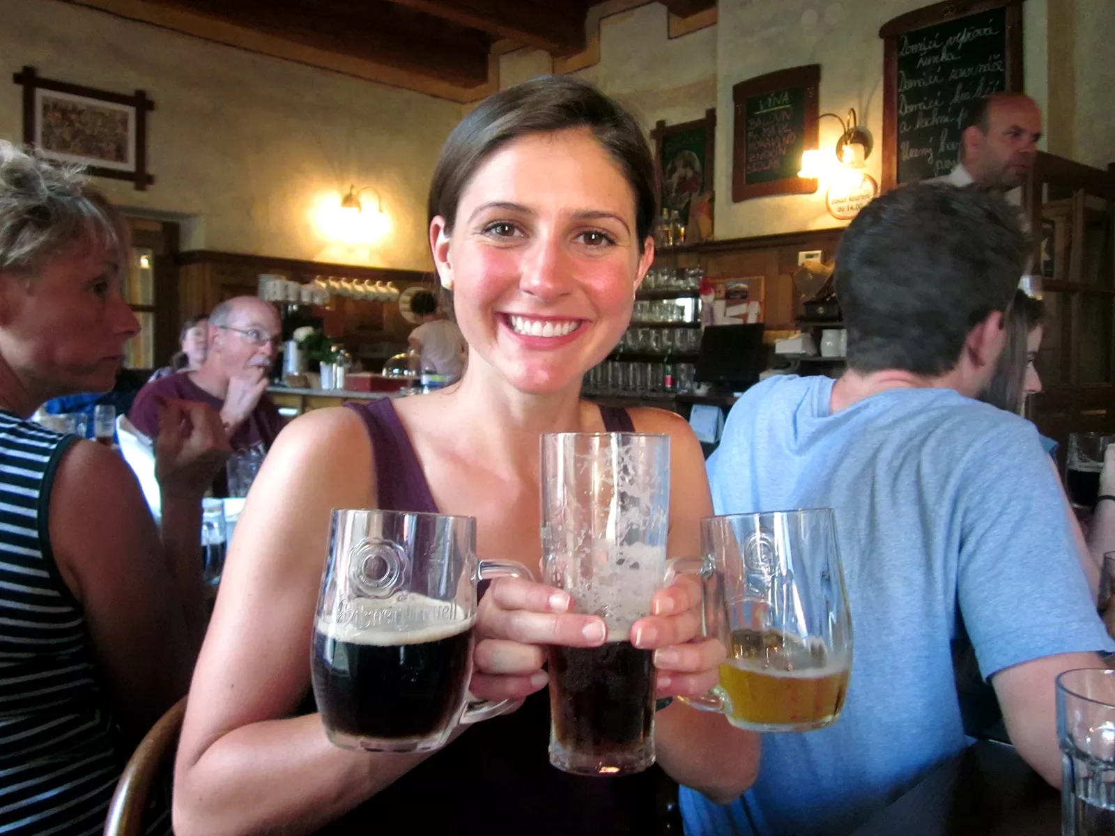 Woman posing in bar with three glasses of beer in varying colors.