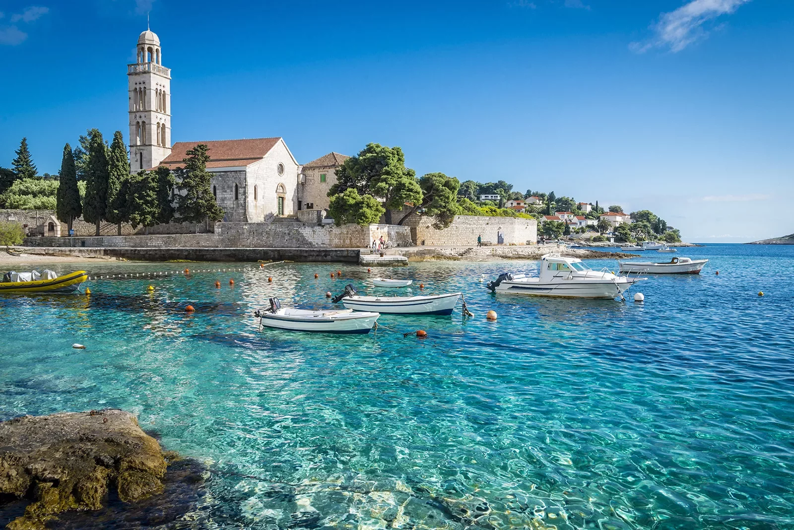 Wide shot of Hvar Island, white stone buildings, blue water, boats.