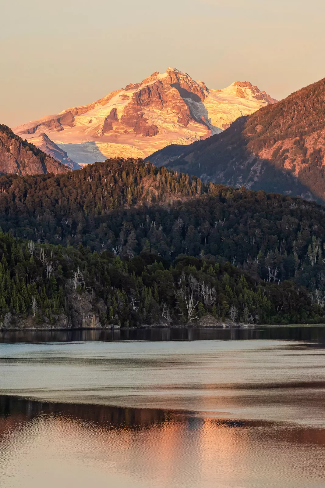 Wide shot of reflective lake during sunset, forest, snowy mountain behind.