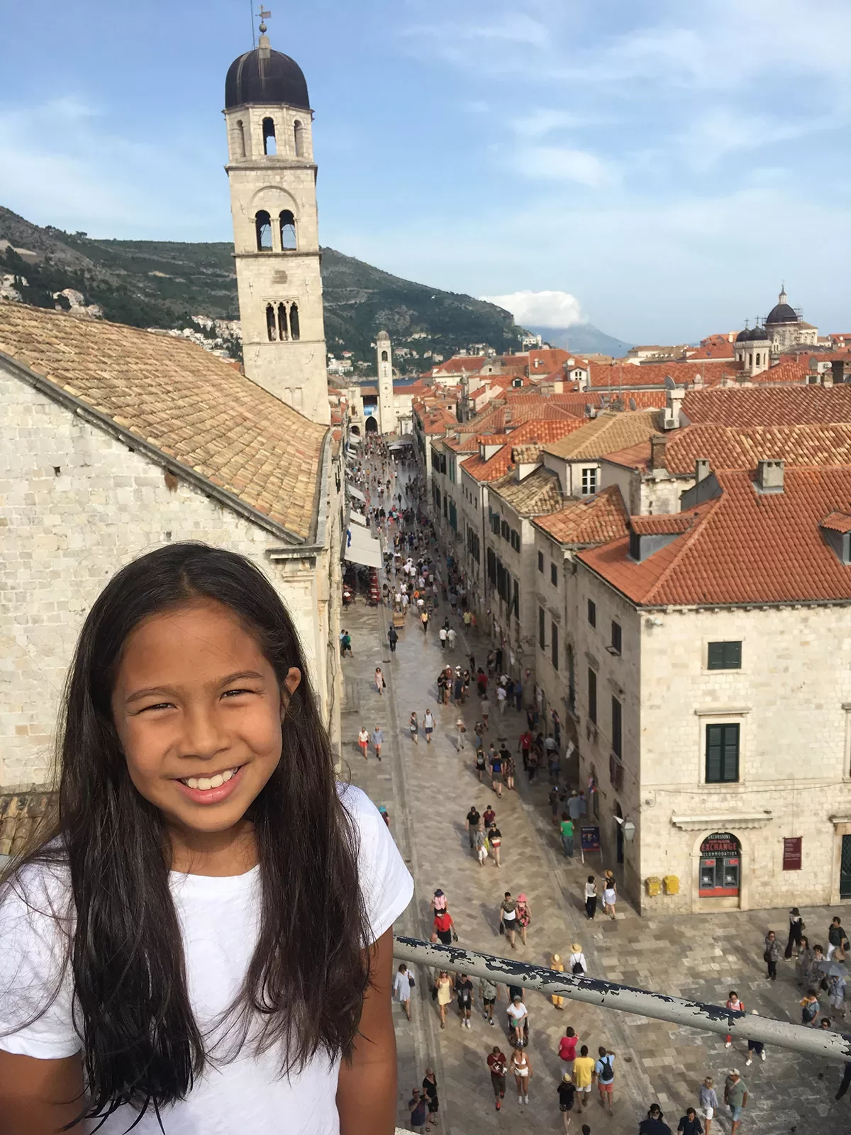 Young guest on balcony, smiling, overlooking crowded Dubrovnik street.