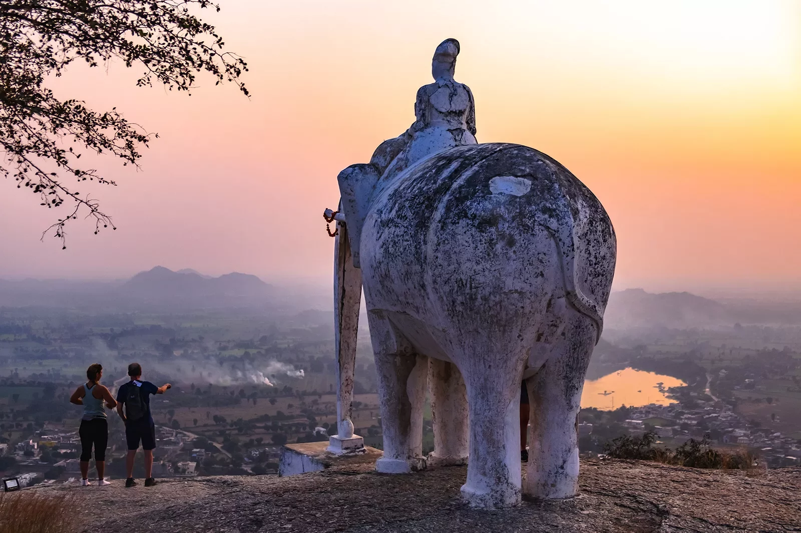 Two people standing beside a large elephant statue as the sun sets in India