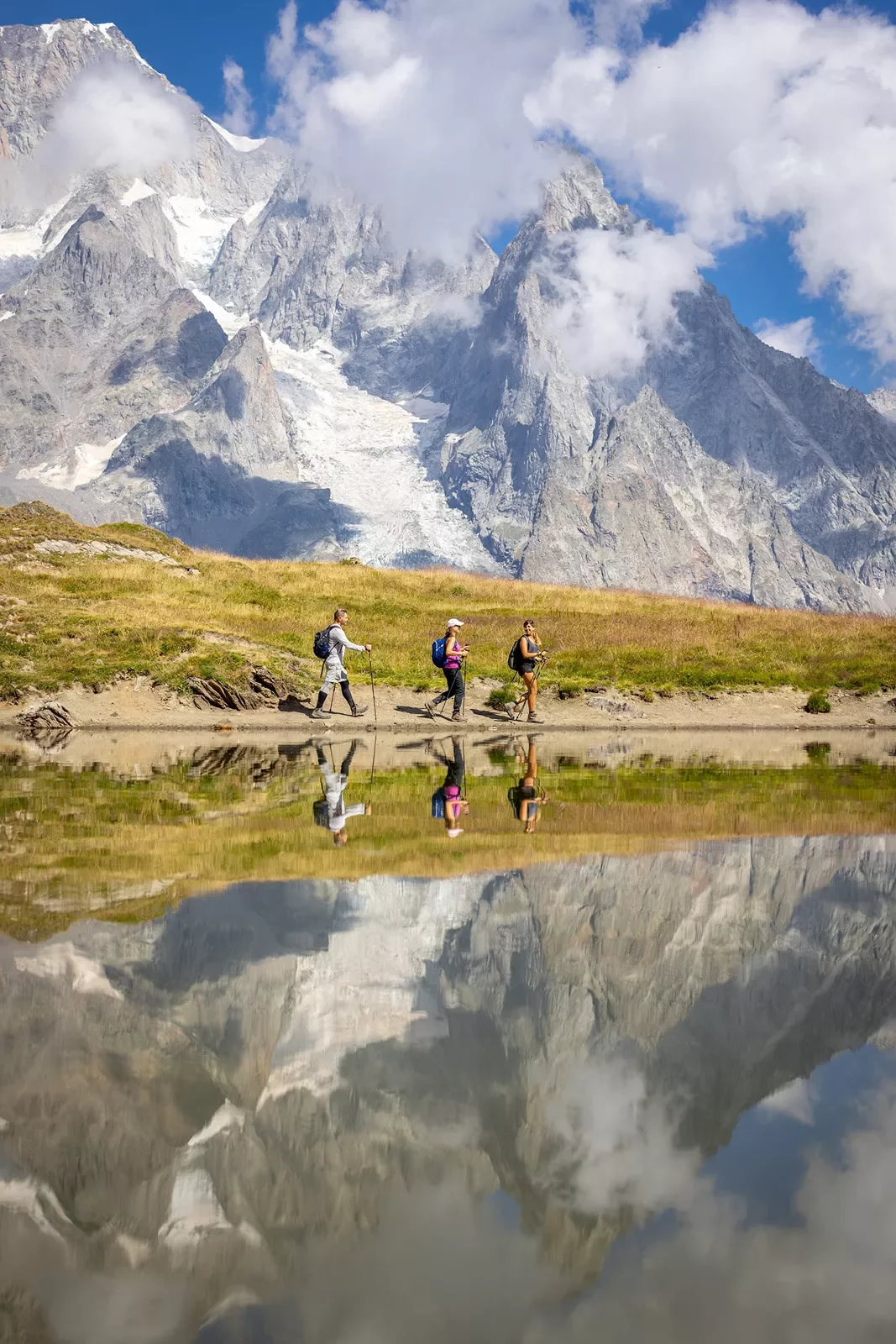 Three guests walking past reflective lake, meadow, snowy mountain in distance.