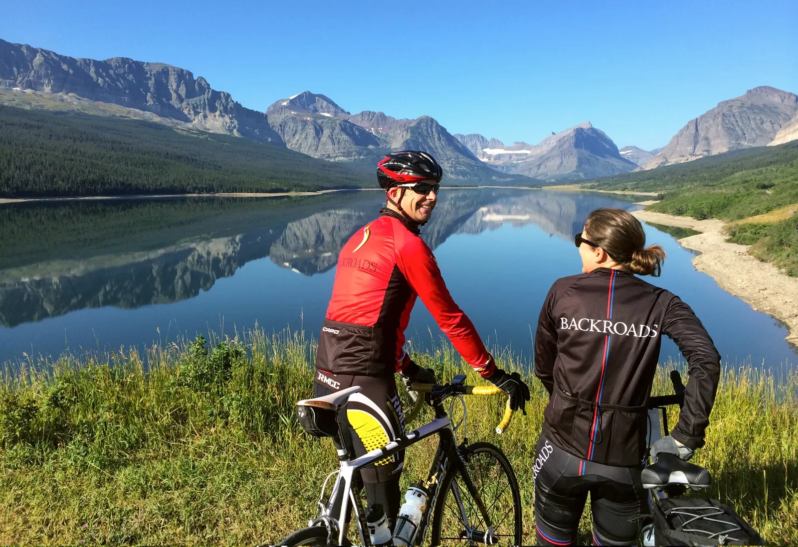 Two bikers resting and chatting with mountains and a clear lake in the background