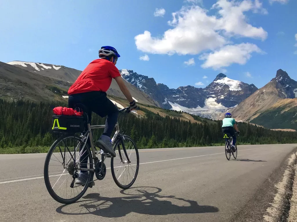 Two guests cycling down mountain road, snowy caps in background.