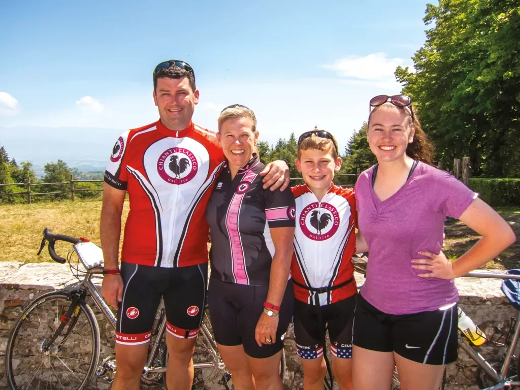 Family in cycling gear poses together in front of bikes and a low wall.