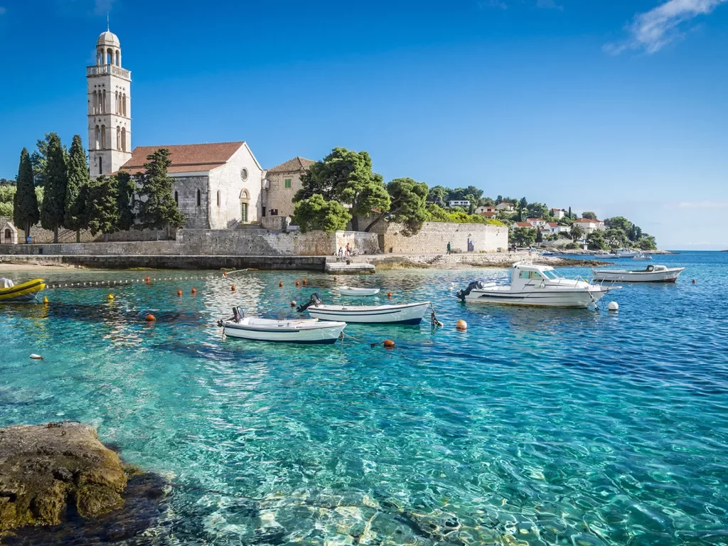 Wide shot of Hvar Island, white stone buildings, blue water, boats.
