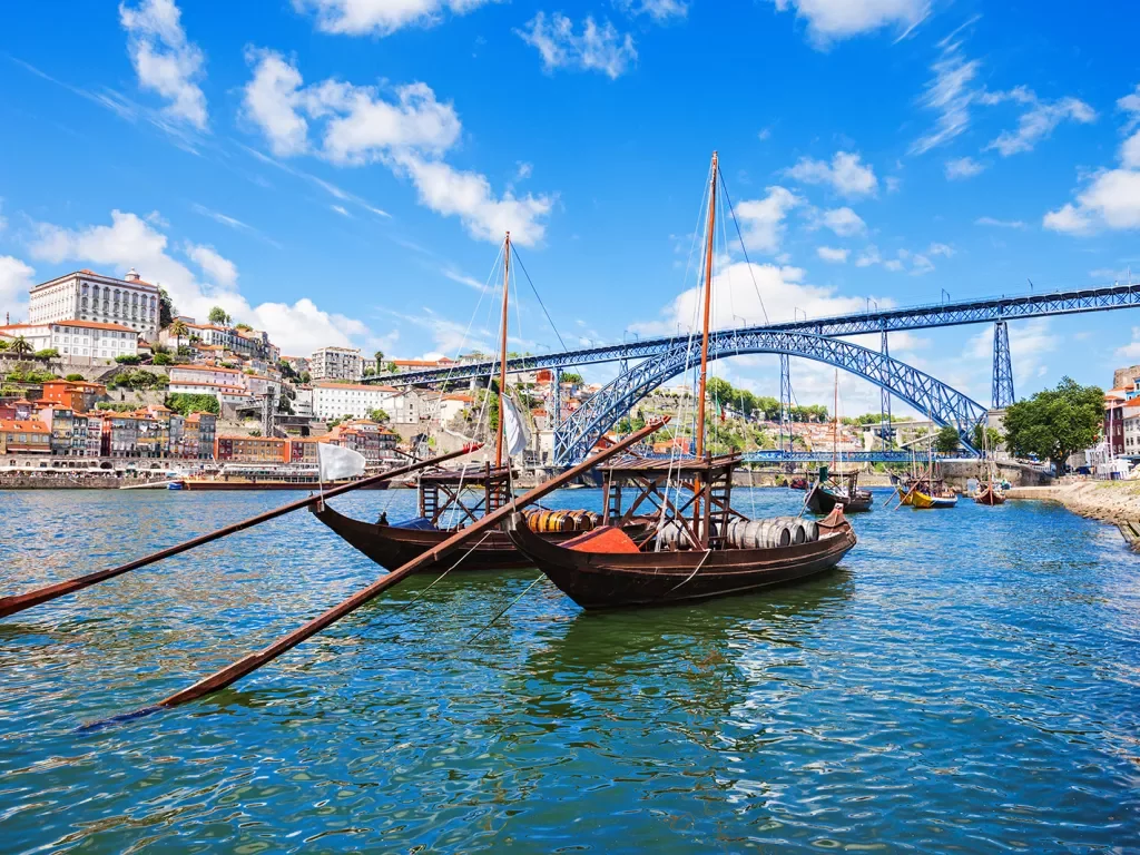 Two dark wood boats on Douro River, large metal bridge, colorful houses behind.