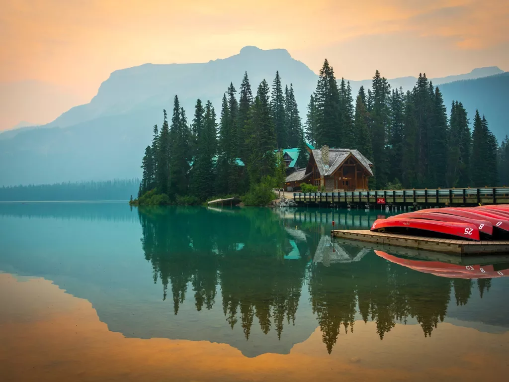 Wide shot of large lake, wooden house, trees, canoes, mountains visible.