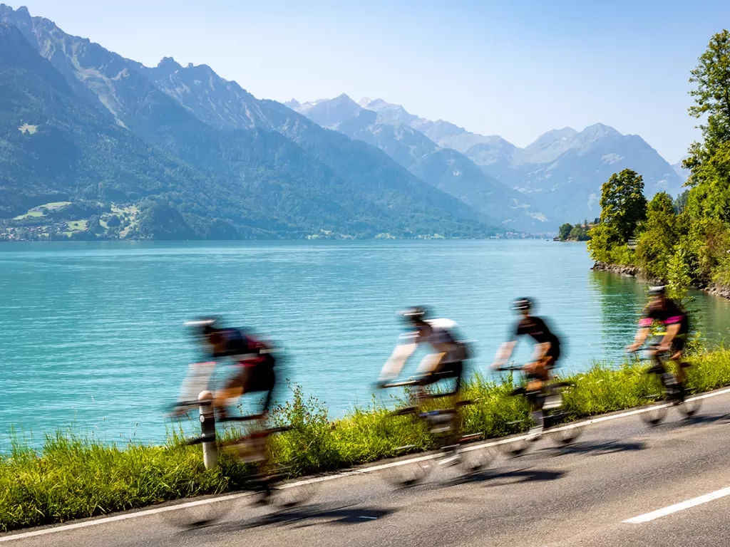 Four guests cycling quickly past blue lakefront, mountains.