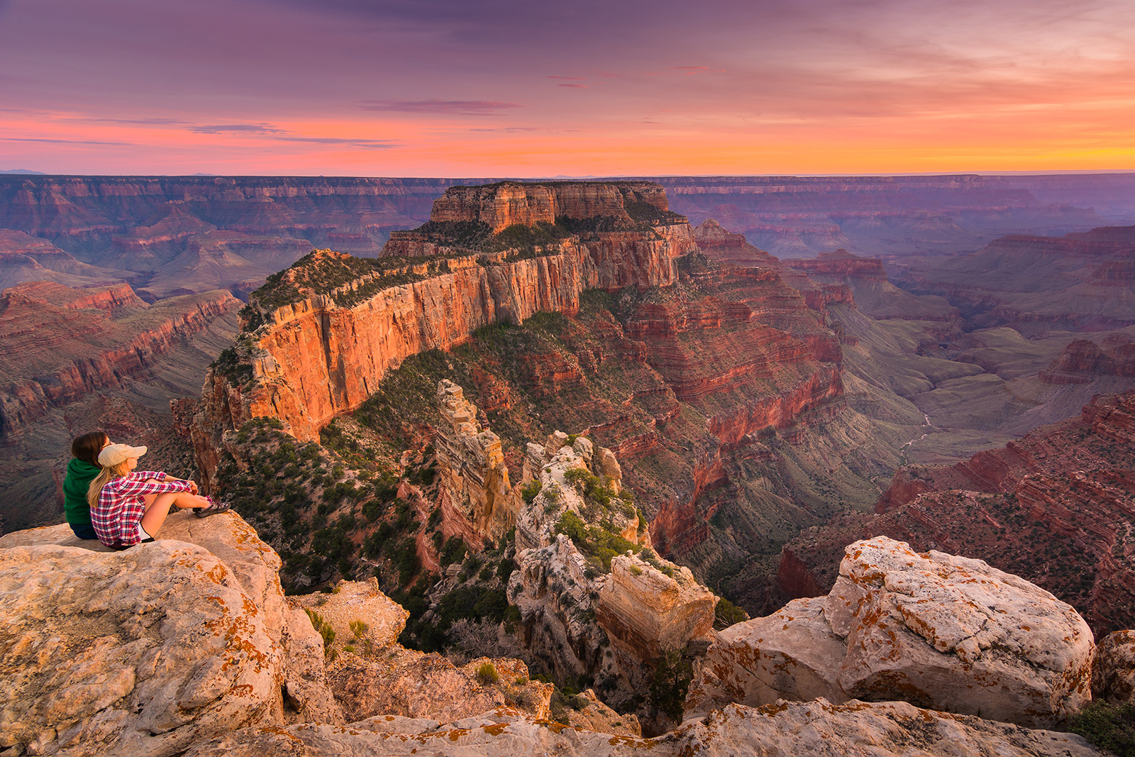 Two guests on cliff, overlooking large orange rock valley during sunset.