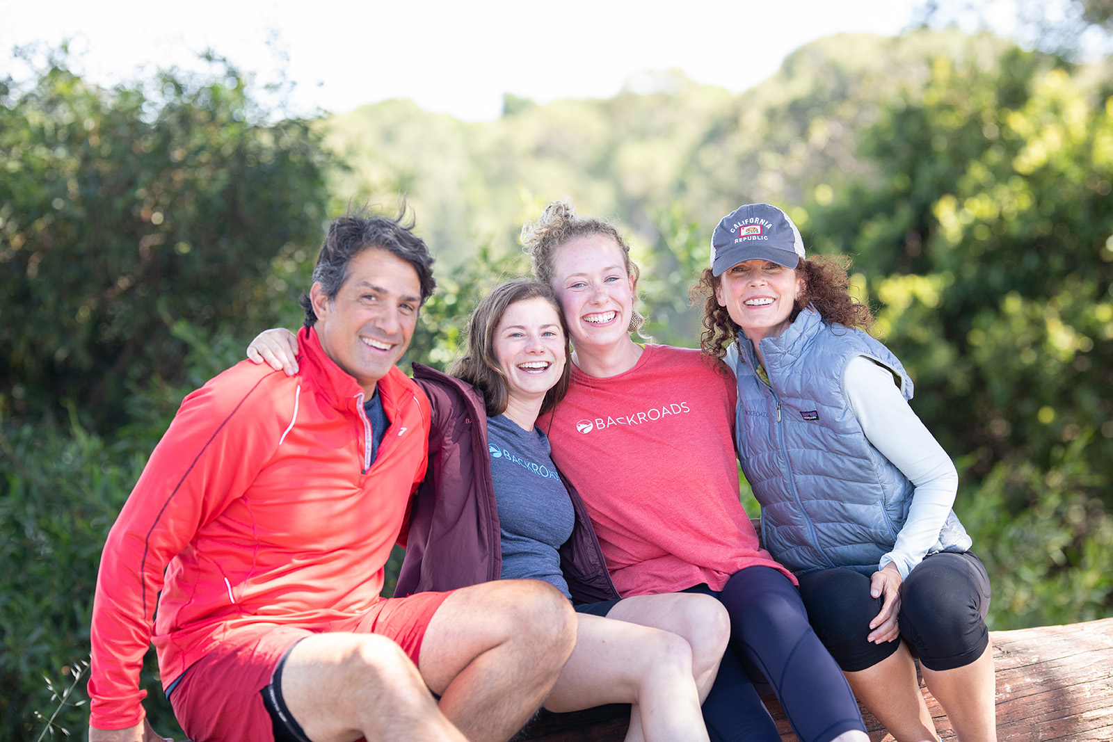 Four guests sitting on log, smiling at camera, forest behind them.
