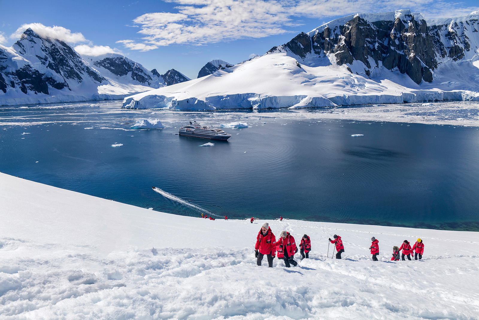 Hikers trekking up a snowy hill in Antarctica with a cruise ship anchored in the background