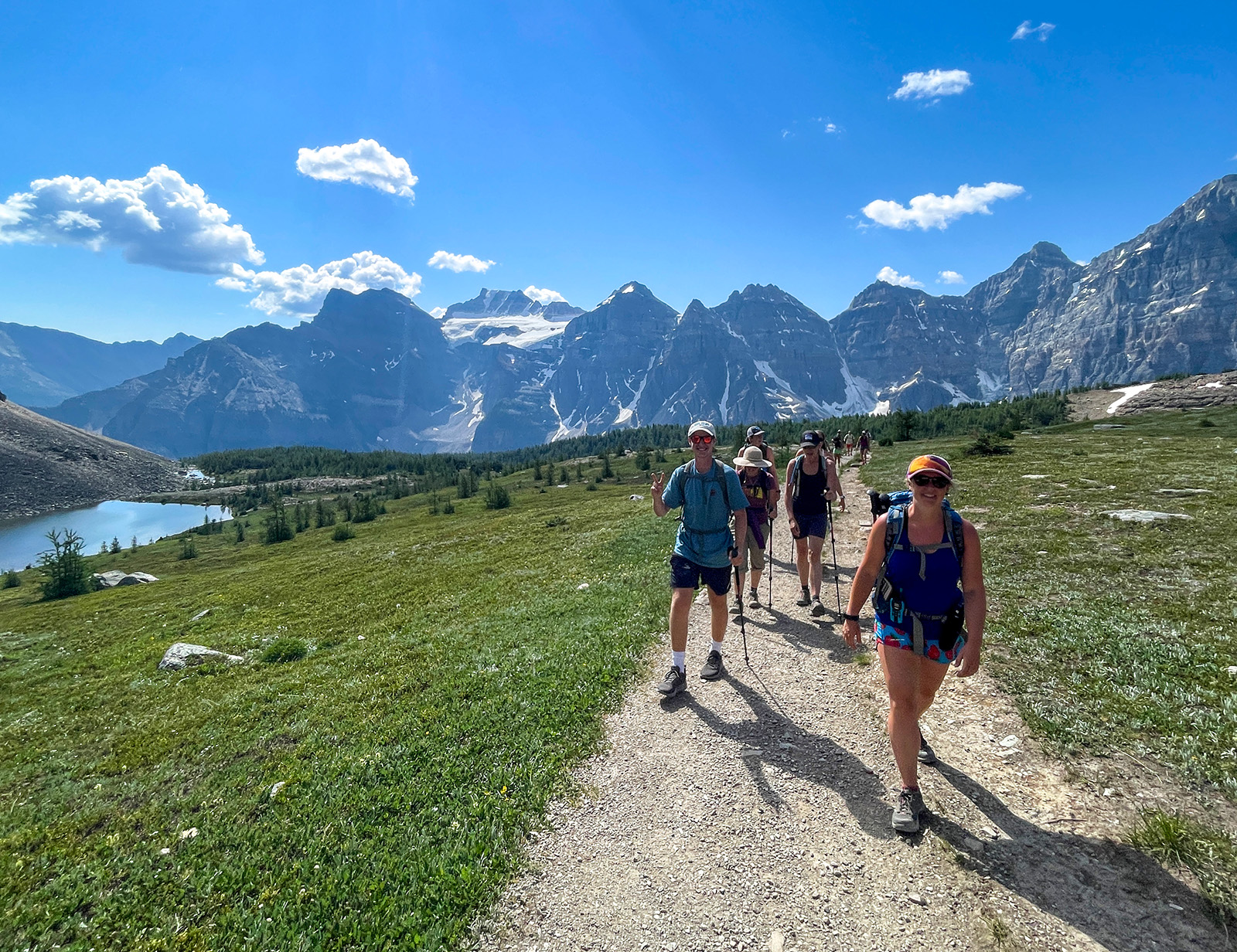 Group of guests walking next to small river, grassy meadow, mountains in background.