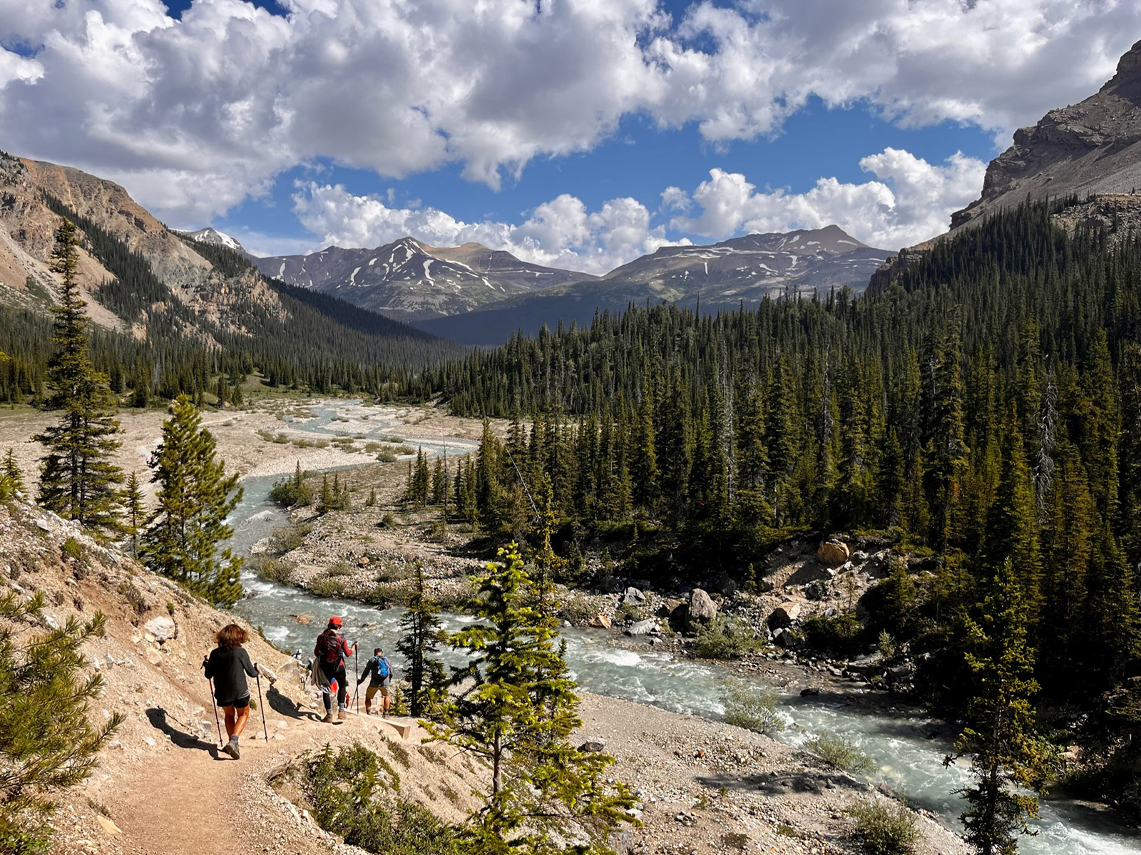 Three guests hiking down hillside, towards flowing stream, mountains, forest.