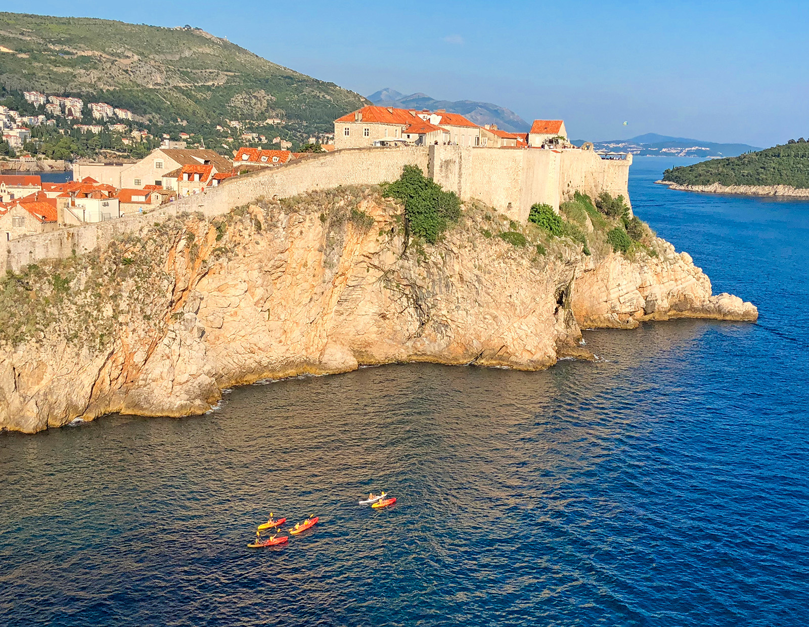 Wide shot of walled cliffside houses, guests kayaking below.