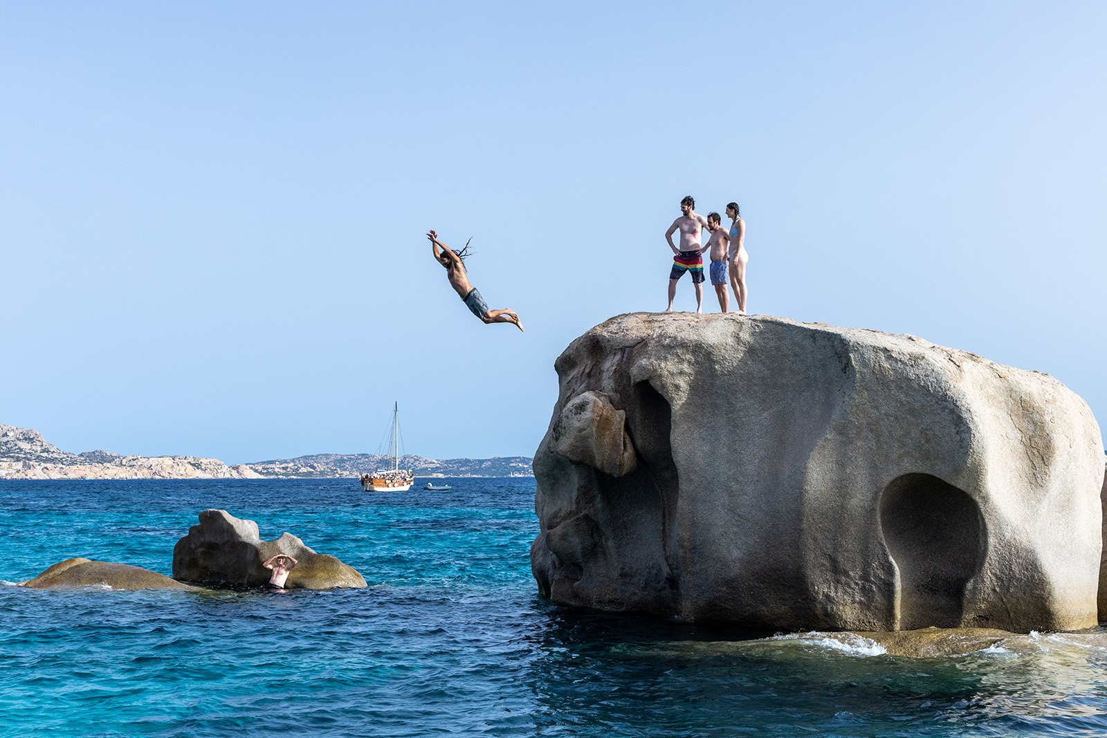 Four guests jumping off large rock, one in the water below.