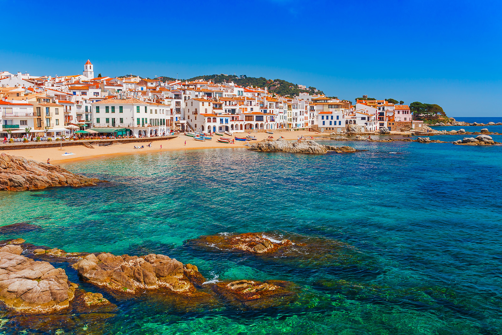 Shot of Costa Brava coastline, blue water, tan and white buildings.