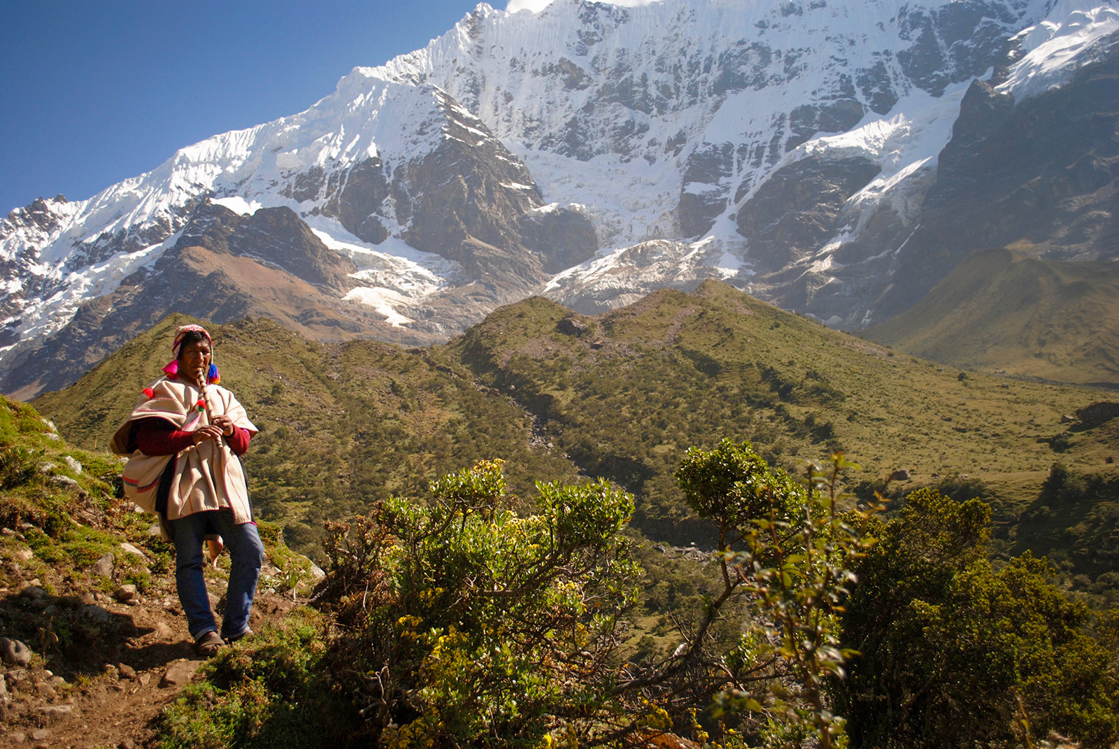 Local on hillside playing instrument, snowy mountains, hills behind them.