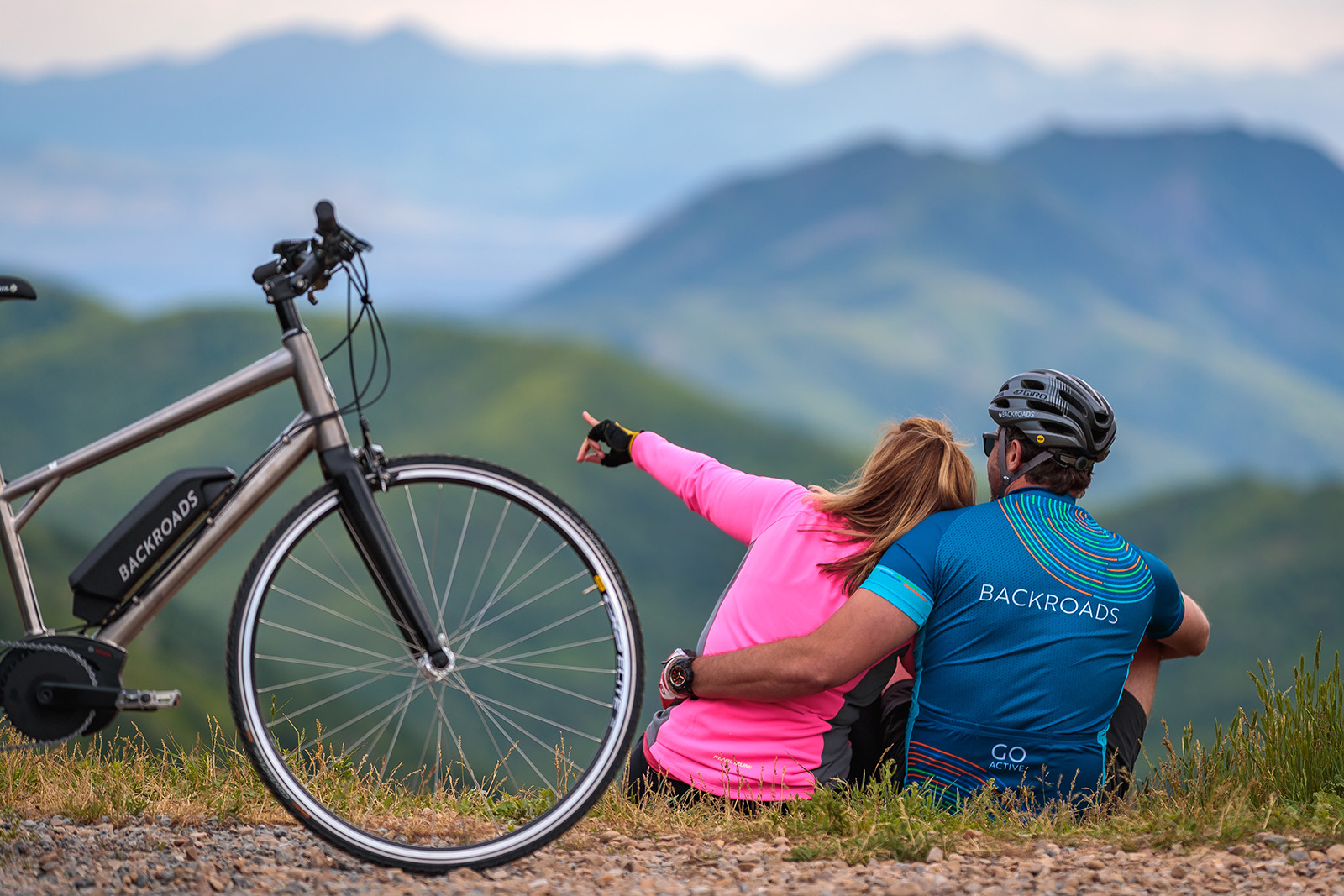Two guests on roadside, pointing towards horizon, bike behind them.