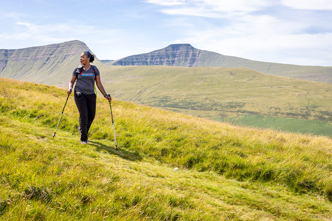 Woman with walking poles descending a tall, grassy will in an open valley