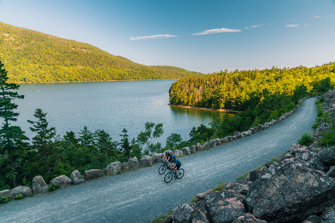 Two bikers on a gravel path, with a lake and tall trees in the distance
