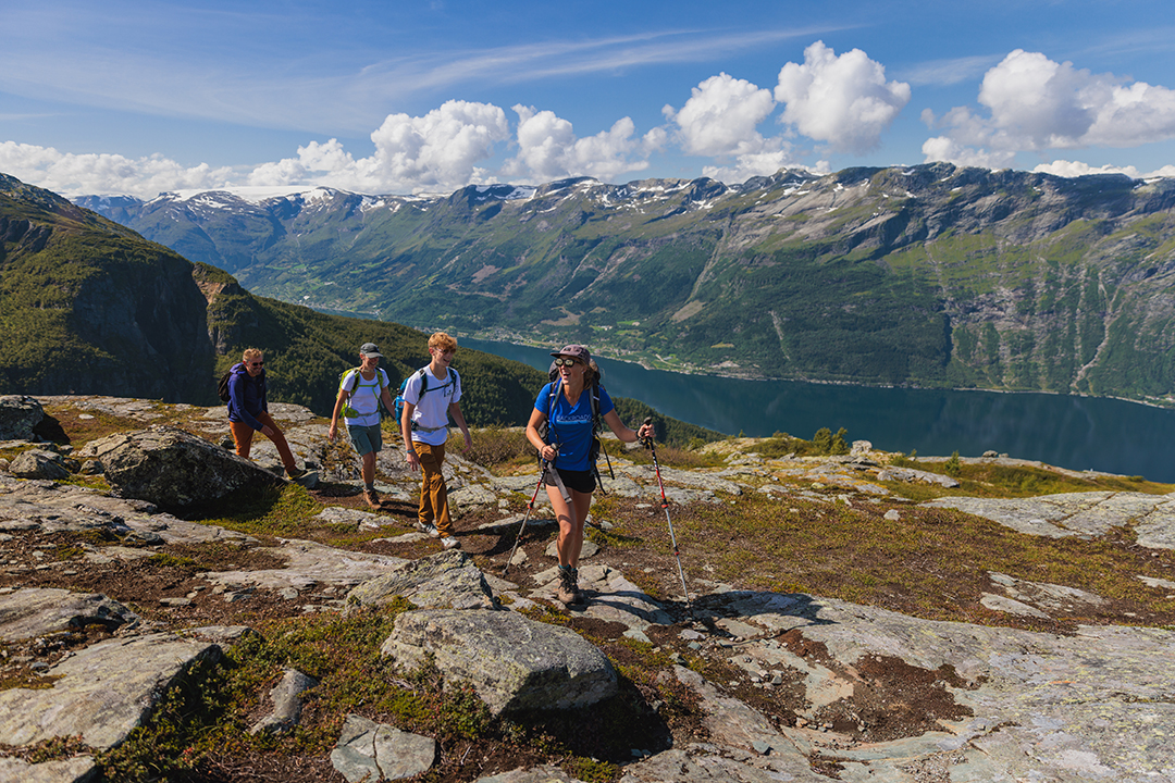 Group of three men and one woman hiking on a dirt and stone trail, with a large lake and mountains in the background