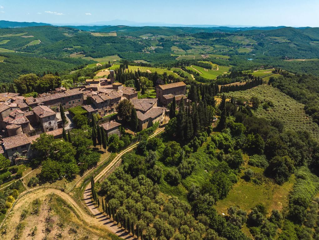 Sky view of brown brick buildings surrounded by a valley of trees and crops
