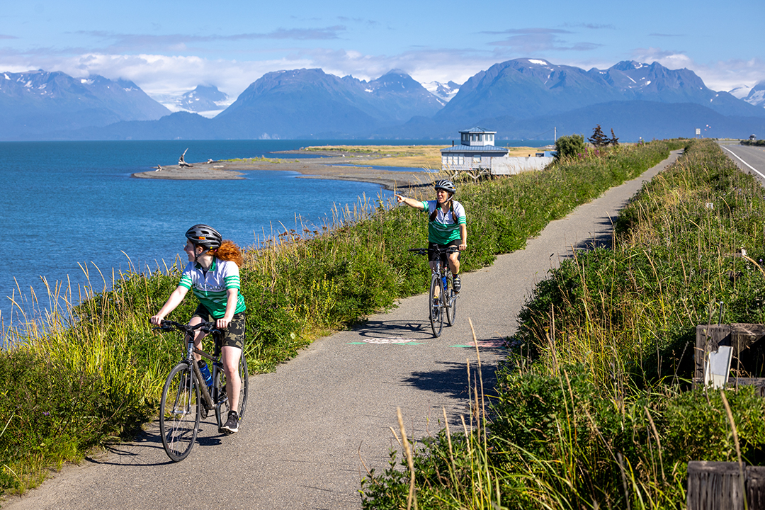 Two women riding bikes along the ocean, pointing towards tall mountains