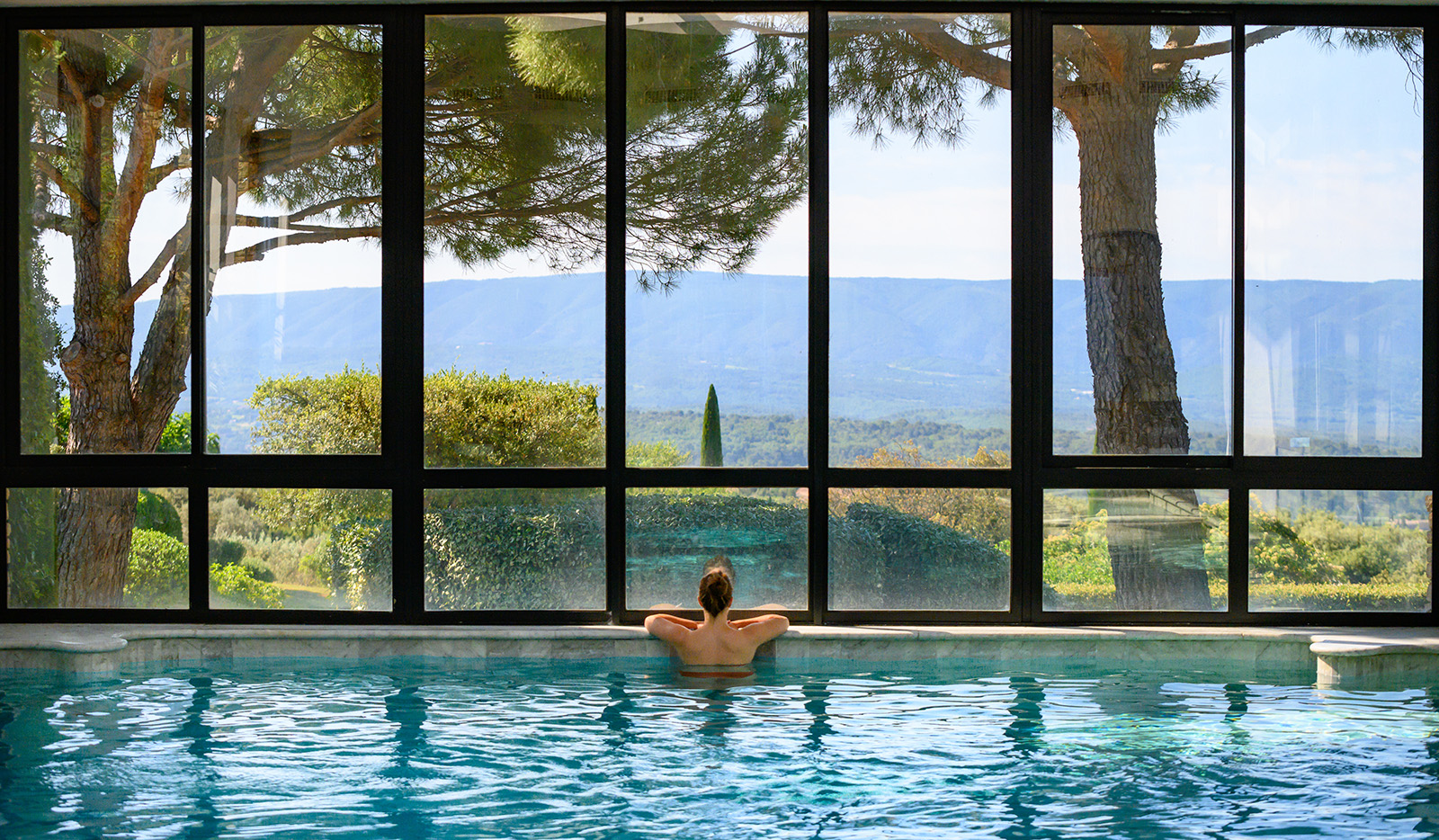 Woman inside of a swimming pool, with her arms on the walkway looking out large windows to a valley