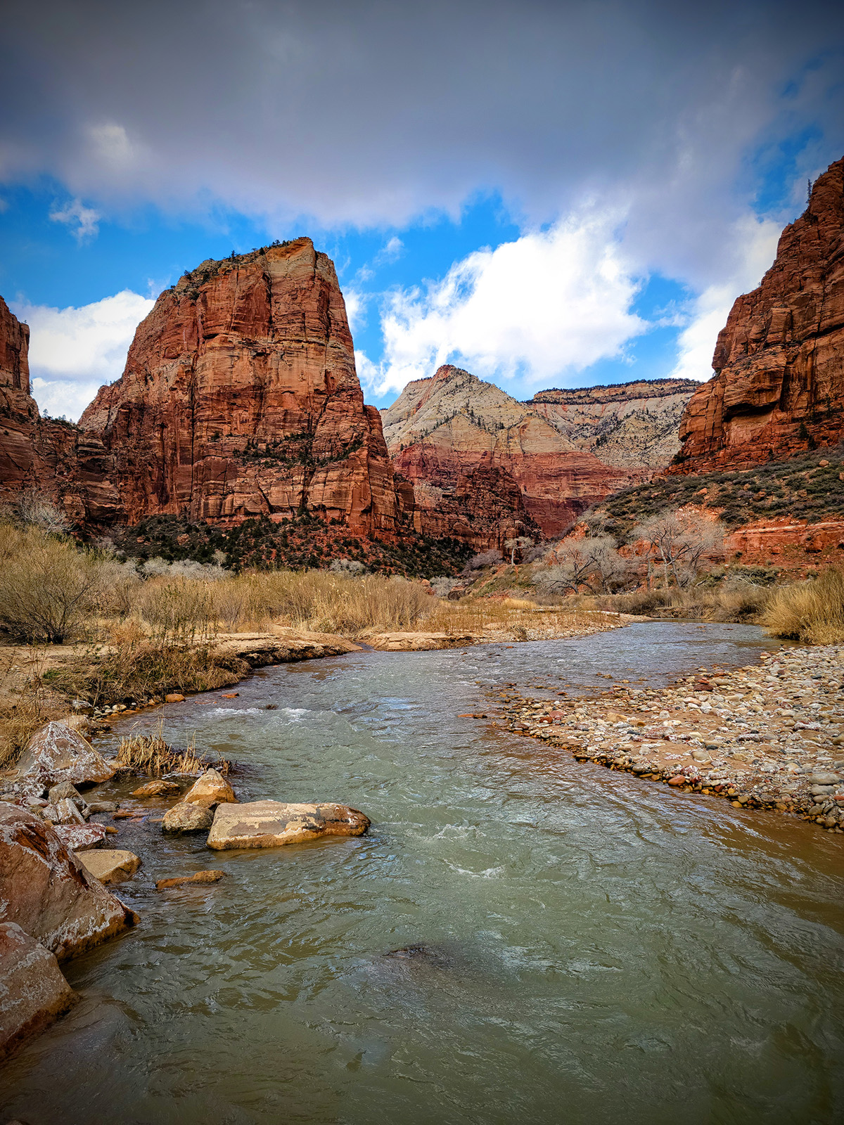 Large, orange canyons with a river on the ground floor, surrounded by gravel and dried plants