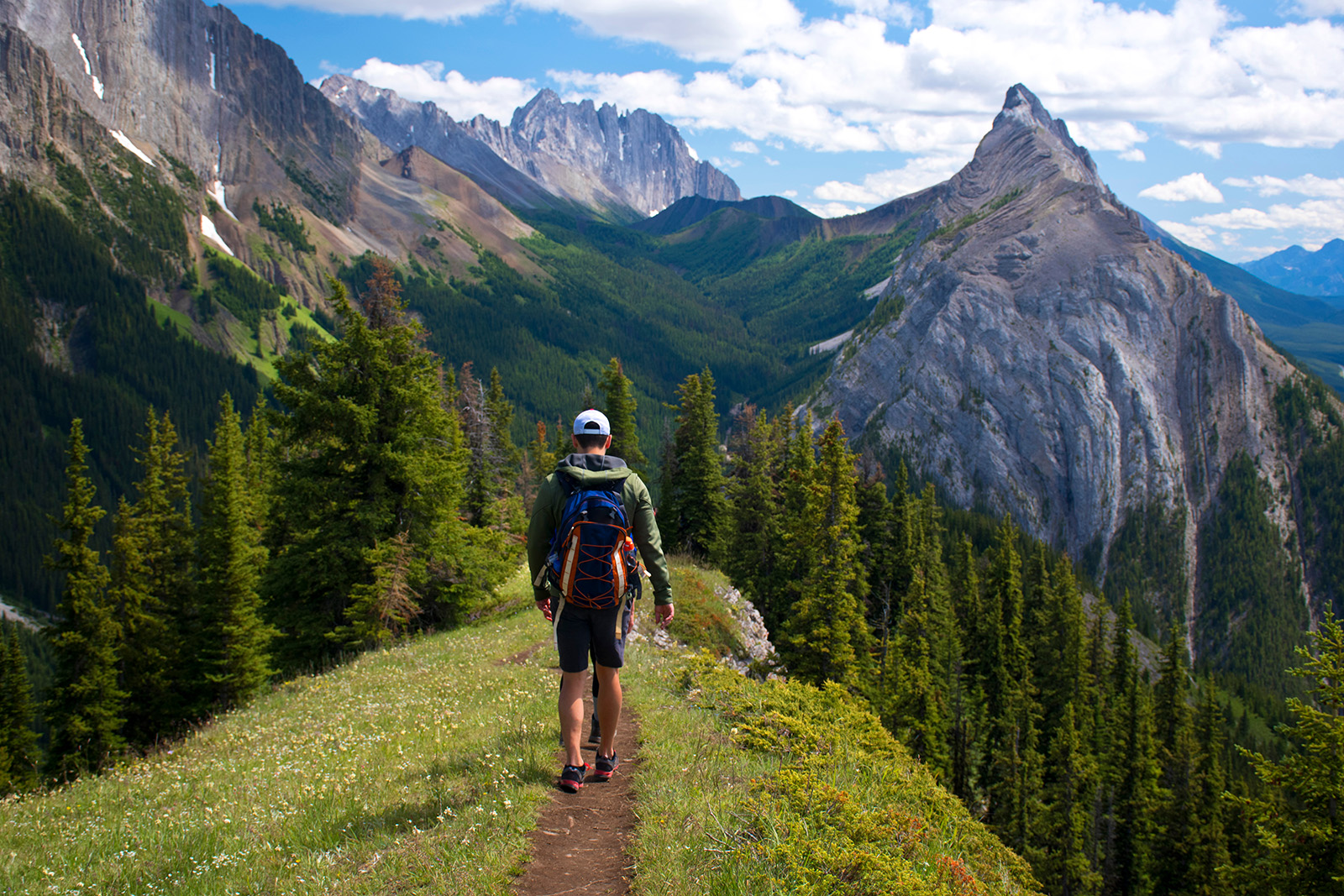 Man walking on a dirt and grass trail towards tall mountains