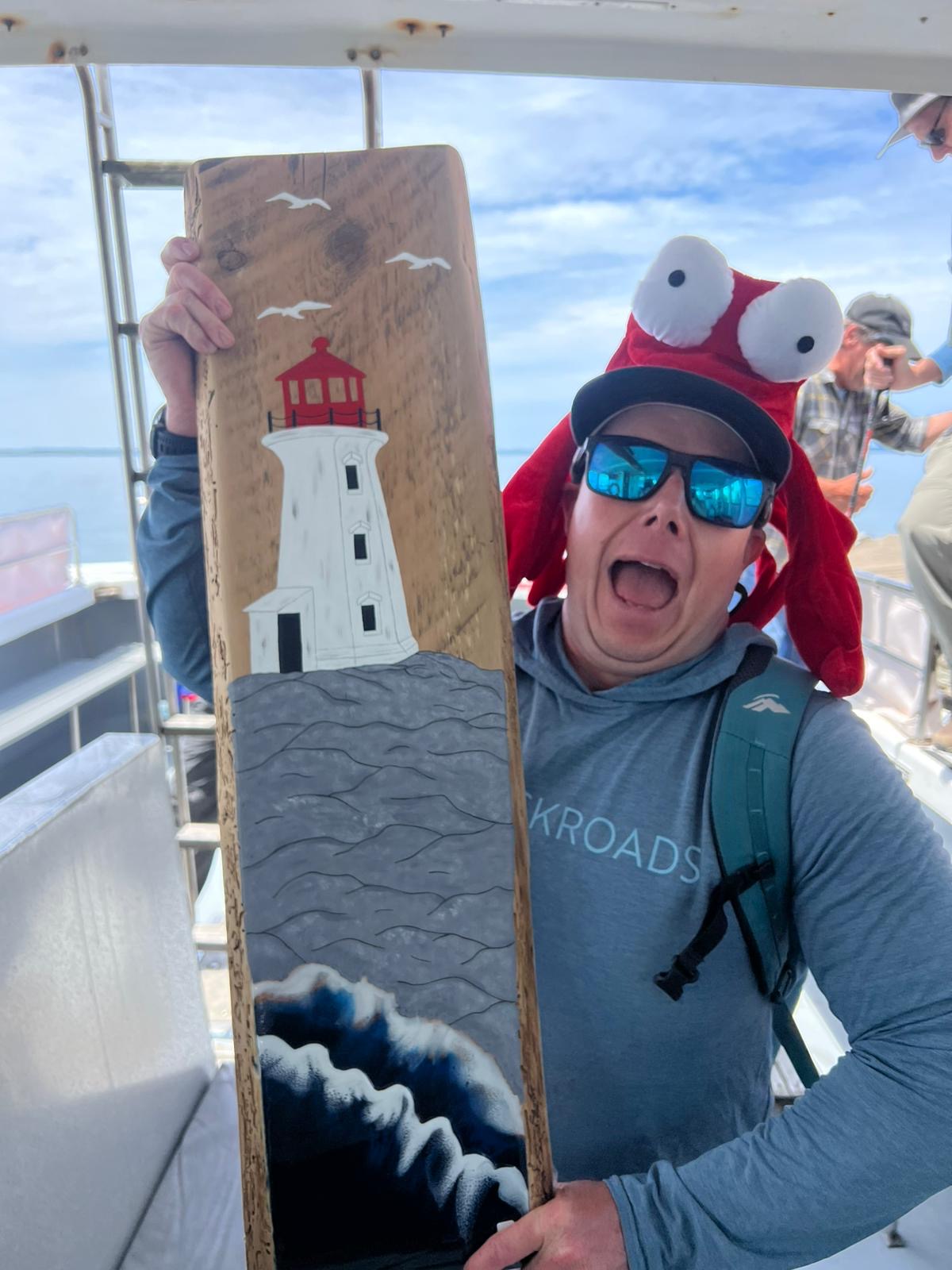 Man wearing a lobster hat, holding up a wooden plank with a lighthouse painted on it
