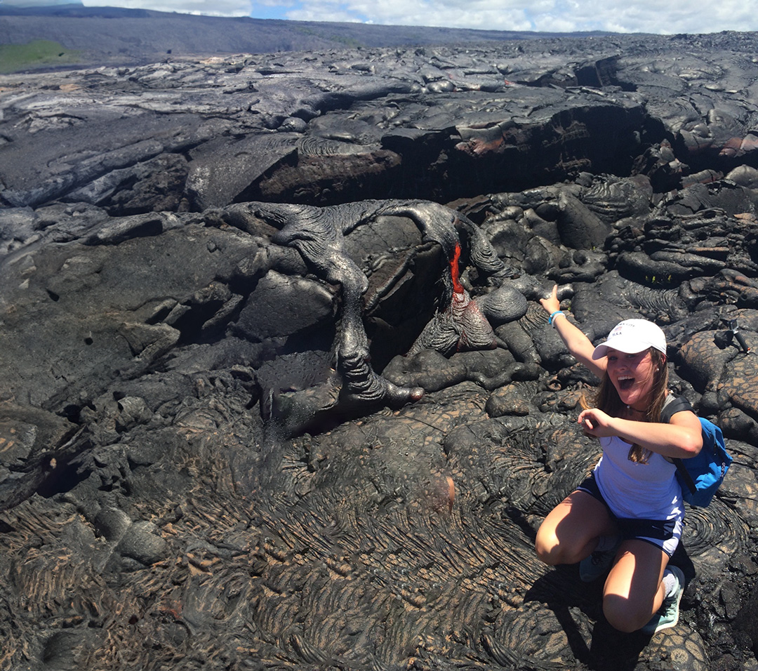Girl wearing a white hate, smiling while pointing at dried lava