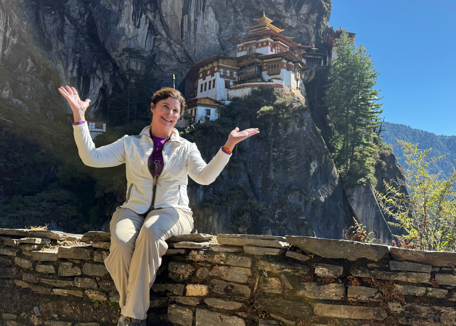 Woman smiling with her arms open, sitting on a stone wall with a temple in the distance