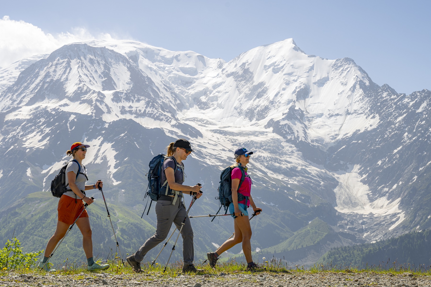 Three women walking on a dirt trail with snow-capped mountains in the background