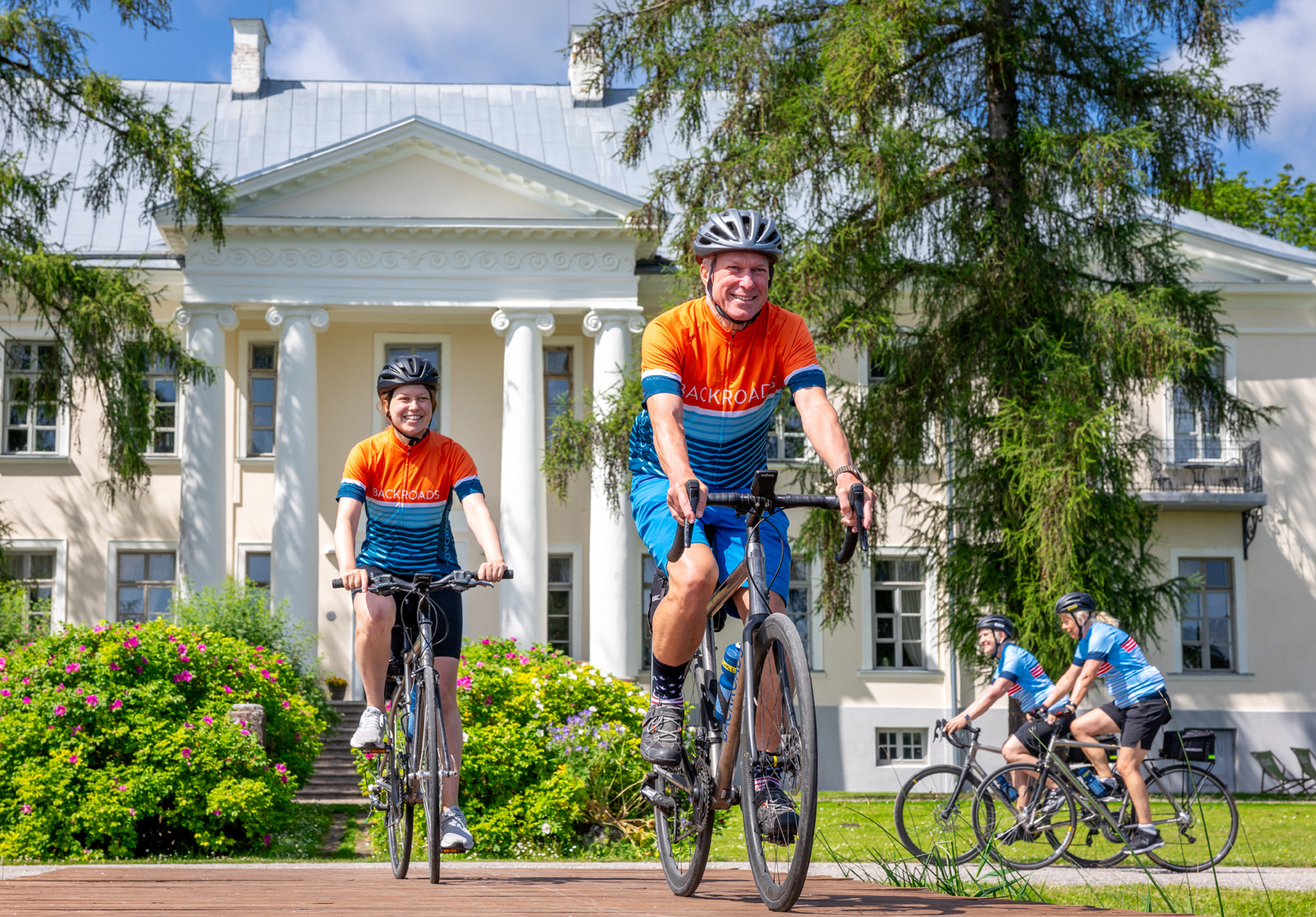 Two couples of men and women smiling while biking in front of a large, white building