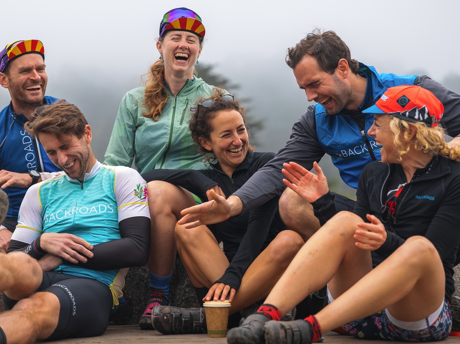 Group of people wearing biking gear, laughing while sitting on a ledge