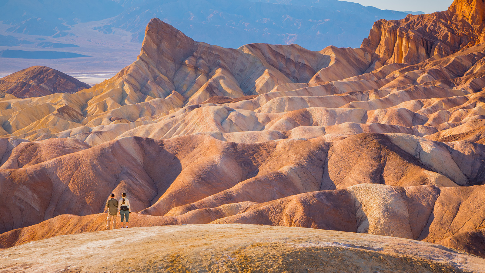 Two people standing on top of a large, orange canyon looking at larger canyons