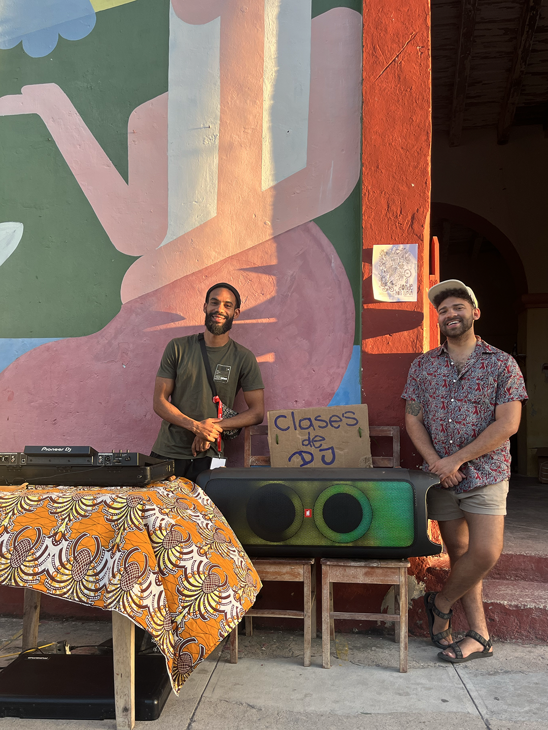 Two men standing by a mural wall with DJ turntables in front