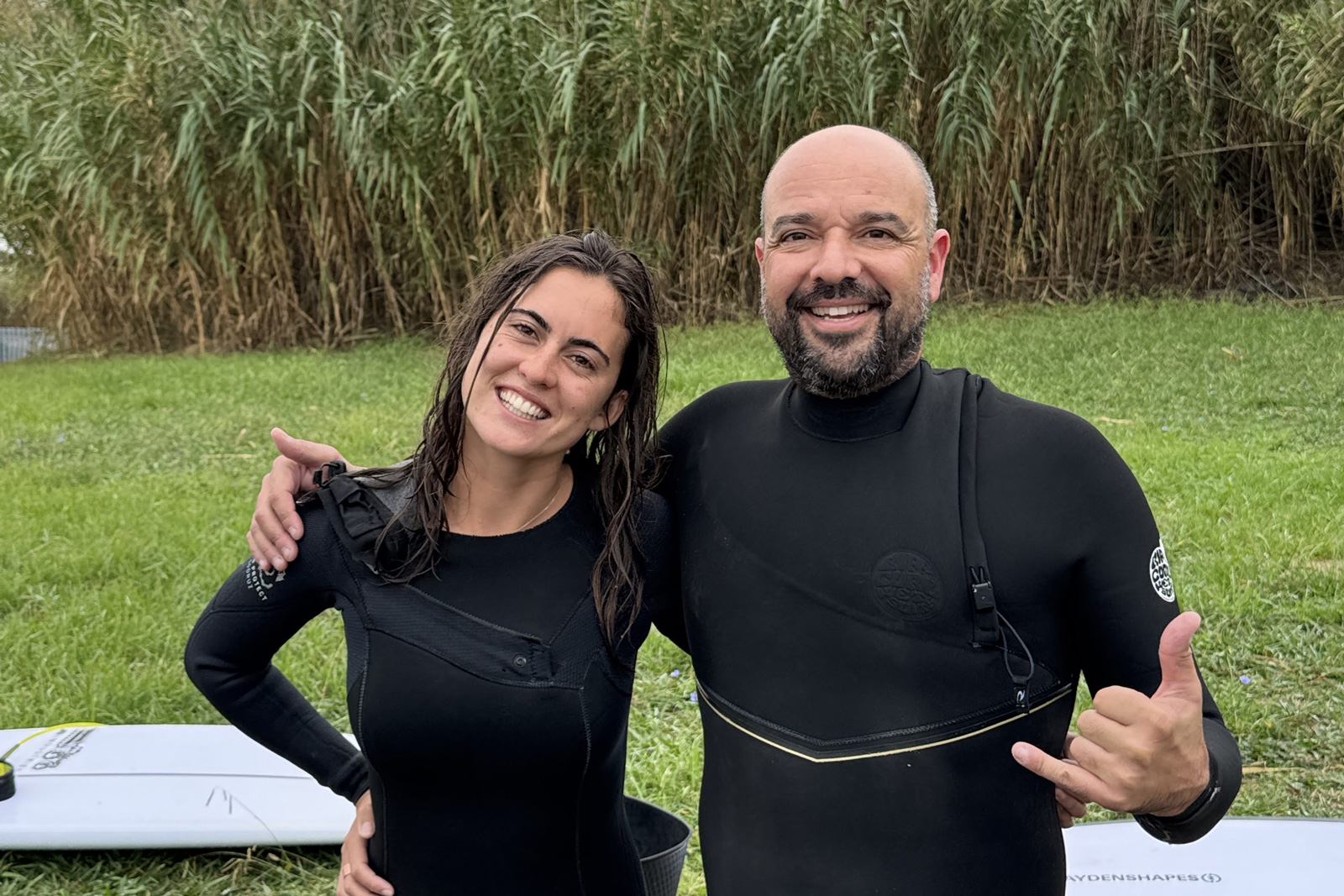 Man and woman in wetsuits, smiling in front of a grass field