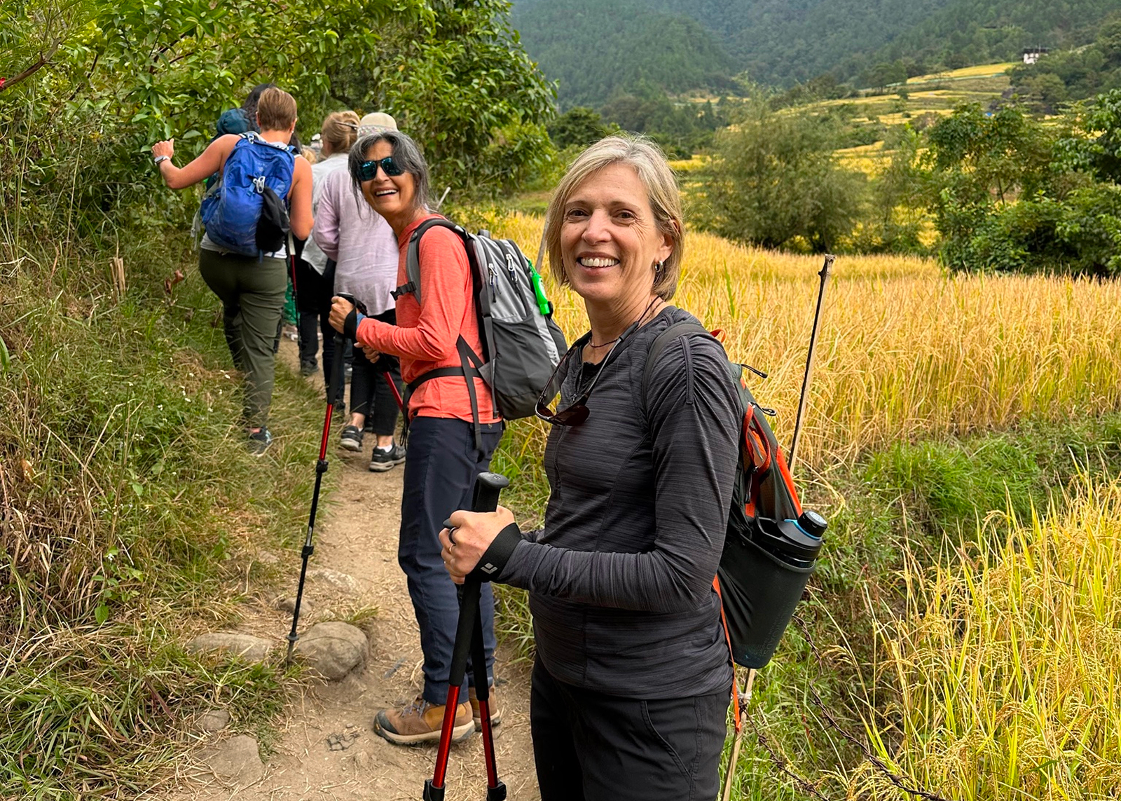 Group of women smiling while hiking on a dirt and weed trail