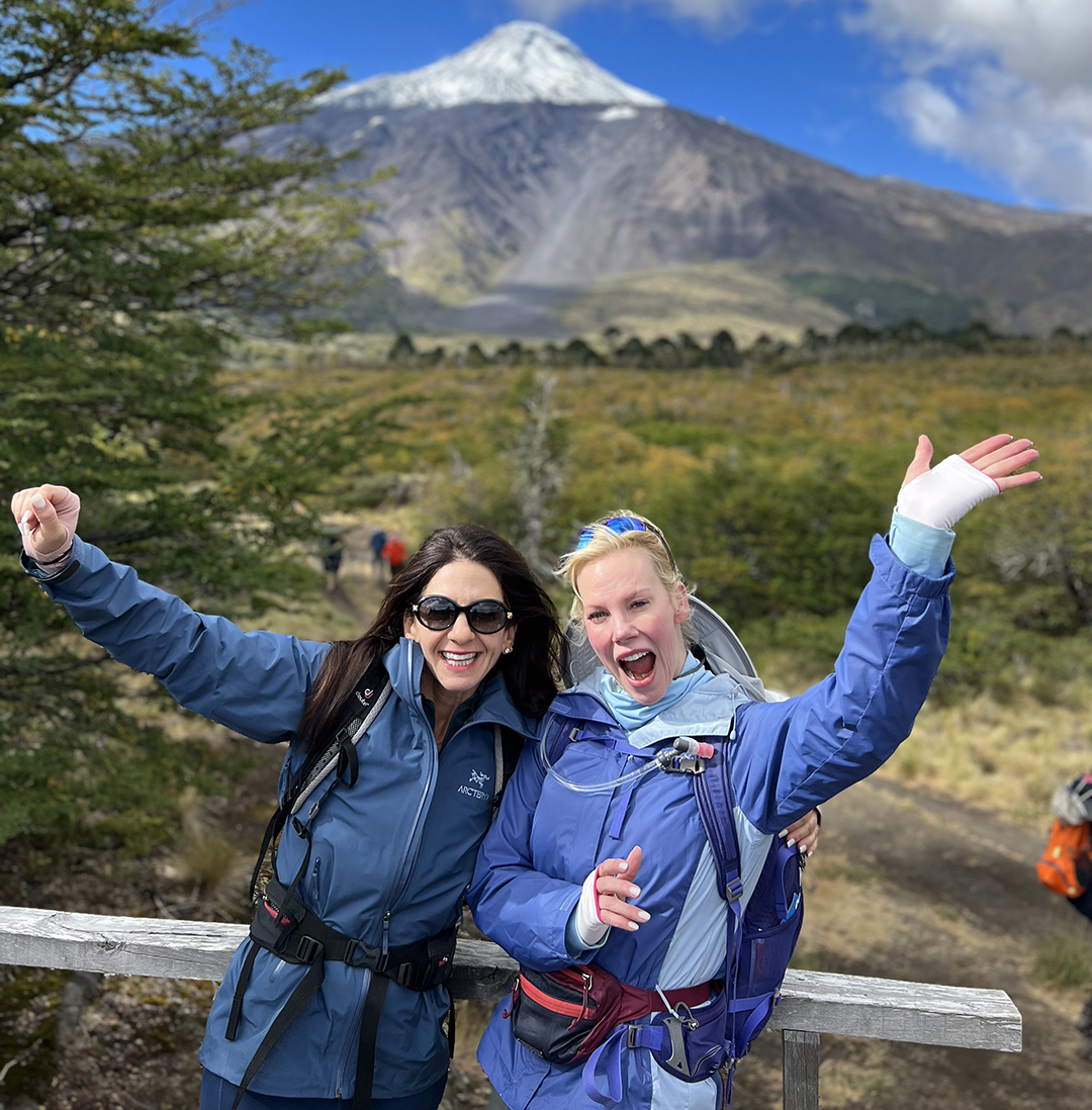 Two women with their arms open and smiling, with a large mountain in the background