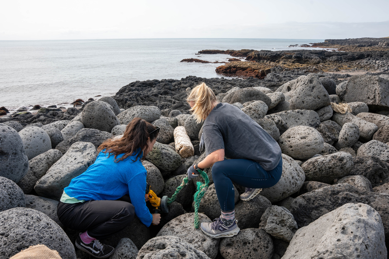 Two women kneeling down by the shore on top of rocks, looking down