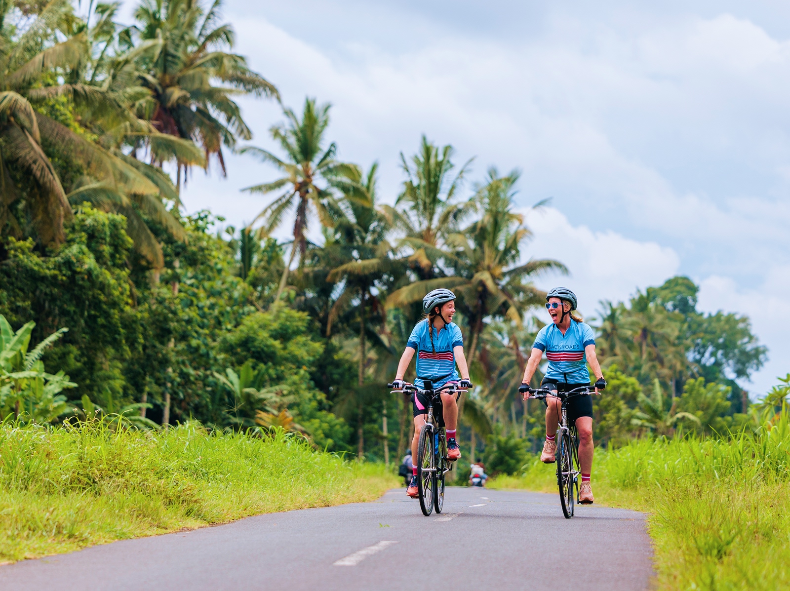 Two people riding bikes on an empty road surrounded by palm trees